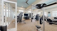 Located at 65 Sentosa Dr in Saint Johns, FL, this fitness center spans a generously sized single-floor layout divided into two dedicated exercise rooms, offering a comprehensive workout environment for all fitness levels. The main exercise room is bright and spacious, featuring large windows along the walls that flood the space with natural light. It is outfitted with a wide array of cardio equipment, including treadmills, ellipticals, stair climbers, and rowing machines, alongside a robust selection of strength-training machines and a dumbbell rack. Full-length mirrors line portions of the walls, enhancing the sense of openness. Ceiling fans with recessed lighting are mounted within tray ceilings accented in soft blue, adding an architectural touch to the otherwise neutral, beige-toned interior. Carpeted flooring throughout the main area provides comfort underfoot during strength sessions. The second exercise room serves as a dedicated studio space, distinguished by its stylish striped hardwood flooring. This room houses a row of stationary cycling bikes equipped with individual screens, a TRX suspension system, medicine balls, and a flat bench, making it ideal for group or individual training. Together, the two rooms create a well-rounded, thoughtfully designed fitness facility with a clean, welcoming ambiance.