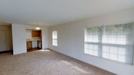 This single-floor space features an open layout that seamlessly connects the living area to the kitchen. The living area is finished with wall-to-wall beige carpet, providing a neutral and comfortable foundation that complements the clean white walls. Two windows fitted with horizontal blinds allow natural light to fill the room, creating a bright and airy atmosphere.

The adjacent kitchen is visible from the living area and features warm-toned wooden cabinetry offering ample storage. Appliances include a white range and a dishwasher, and the kitchen is equipped with a pendant light fixture that hangs above, adding a functional yet decorative element. The kitchen flooring transitions to a harder surface, contrasting with the carpeted living space.

The overall design of the space is straightforward and practical, with a simple, clean aesthetic throughout. The open connection between the living area and kitchen promotes a functional flow, making the space well-suited for everyday living. The abundance of natural light from multiple windows enhances the sense of openness across the layout.