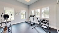 Located in Homestead, FL, this dedicated fitness room offers a clean, modern environment designed to support a well-rounded exercise routine. The space features soaring ceilings that lend an open, airy feel, enhancing the overall sense of room to move and train comfortably. Neutral gray walls and a durable speckled rubber flooring provide a practical yet polished aesthetic throughout.

Large windows line multiple walls, flooding the room with abundant natural light while framing views of the lush tropical surroundings, including swaying palm trees and open green spaces — a refreshing backdrop for any workout. Full-length mirrors are strategically mounted along the walls, visually expanding the space and allowing users to monitor their form during exercise.

The room is equipped with a treadmill, elliptical trainers, and a stationary bike for cardiovascular training, alongside a weight bench and a rack of graduated dumbbells for strength work. A glass-paned white door provides access, contributing to the bright and welcoming atmosphere. The overall design is functional and well-organized, striking a balance between a purpose-built workout environment and a light-filled, inviting interior that motivates an active lifestyle.