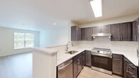 This single-floor, 3-bedroom, 1-bathroom residence offers a thoughtfully laid-out floor plan that maximizes every square foot of living space. The open-concept kitchen is a standout feature, showcasing dark espresso cabinetry with sleek hardware, pristine white quartz countertops, a herringbone tile backsplash, and a full suite of stainless steel appliances including a dishwasher, range, and built-in range hood. A peninsula with an undermount sink provides additional prep space and connects seamlessly to the living area, which is bathed in natural light from a large window and features light-toned hardwood-style flooring. The three bedrooms offer comfortable proportions, with soft carpeting underfoot and windows overlooking greenery. The primary bedroom includes a walk-in closet outfitted with wire shelving. One bedroom features hardwood-style flooring and a built-in closet. The shared bathroom is cleanly appointed with floor-to-ceiling subway tile in the tub/shower surround accented by neutral stripe banding, a dark vanity with a white countertop, and tile flooring. A hallway connects the living spaces efficiently, completing this functional and modern residence.