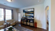 This single-story space showcases a well-appointed living area that blends contemporary design with warm, inviting elements. The room features rich, dark hardwood flooring that contrasts elegantly with the crisp white walls and classic crown molding that frames the ceiling, lending a polished, refined feel to the space.

A striking large-scale landscape painting in deep blues, golds, and whites serves as the room's focal point, displayed prominently above a rustic yet industrial-style cabinet. The cabinet, featuring a combination of dark metal framing and natural wood panel doors, provides both storage and display space, and is styled with decorative sculptures, vases, and accent pieces that add personality and depth.

Natural light filters into the room through a window fitted with white plantation shutters, contributing to the bright and airy atmosphere. Tall decorative floor vases flank the cabinet, adding vertical interest to the composition. A plush area rug anchors the seating area, where a neutral-toned armchair adds comfort. The overall ambiance is modern and sophisticated, with thoughtful décor choices that create a cohesive and welcoming environment.