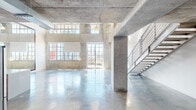 Loft 1013 at 1601 N Miami Ave is a striking two-story industrial loft in the heart of Miami, offering an authentic raw aesthetic defined by exposed concrete ceilings, structural columns, and polished concrete floors throughout. The main level features a dramatically tall, double-height open living area bathed in natural light from oversized multi-pane industrial windows that frame sweeping city views. A sleek stainless steel staircase with modern horizontal railings connects the two floors, adding a refined contrast to the raw surroundings. The kitchen is outfitted with warm wood cabinetry, stainless steel appliances including a large French door refrigerator, a built-in oven, and an integrated range hood, all set against the exposed concrete backdrop. The upper level offers an open loft area with polished concrete floors, exposed mechanical piping highlighted in red, and access to additional rooms. A well-appointed full bathroom features a double vanity with wood cabinetry, a glass-enclosed shower, and an industrial-style window overlooking the Miami skyline. A secondary utility space includes a half bath and in-unit stacked washer and dryer. Throughout, exposed ductwork, red fire suppression pipes, and raw concrete surfaces reinforce the space's bold industrial character.