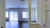 This single-floor space showcases a clean, modern interior designed to maximize openness and natural light. The main living area features rich, dark hardwood flooring that contrasts elegantly with the bright white walls and ceiling, creating a crisp, contemporary aesthetic. Large windows fitted with horizontal blinds line the walls, while a full-height sliding glass door dressed with vertical blinds floods the space with abundant natural light and offers views of surrounding greenery outdoors. The open layout is defined by a structural column and a partial wall with a built-in ledge, which subtly divides areas while maintaining an airy, connected feel throughout. Ceiling-mounted HVAC vents indicate a centralized climate control system integrated seamlessly into the design. The neutral color palette of soft whites and grays, combined with the warm tones of the hardwood flooring, lends the space a versatile and welcoming ambiance. The absence of furnishings allows the architectural details — including clean lines, smooth walls, and consistent flooring — to take center stage, highlighting the thoughtful layout and practical design of this well-lit, open interior.