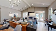 This single-story clubhouse lounge is defined by its soaring vaulted ceilings, which lend the space an open, airy quality throughout. A dramatic multi-ring chandelier serves as the room's centerpiece, casting warm light across the generously proportioned main gathering area. Light-toned large-format tile flooring runs throughout, contributing to the bright, cohesive aesthetic.

The main lounge area is anchored by a sleek linear fireplace mounted below a wall-mounted television, flanked by French doors that flood the interior with natural light and provide direct access to an outdoor area. Comfortable seating arrangements — including a tufted cognac leather sofa, upholstered armchairs, and vivid blue accent chairs — are organized around colorful area rugs, creating distinct yet connected conversation zones.

Rounded wood bistro tables with contemporary chairs offer casual seating toward the front of the space, ideal for smaller gatherings. A dedicated wet bar area features dark wood cabinetry, a metallic subway tile backsplash, white countertops, a sink, and a built-in beverage refrigerator. Curated artwork, decorative mirrors, and carefully selected accessories add personality throughout. Large windows bring in additional natural light and offer views of the surrounding outdoor landscape.