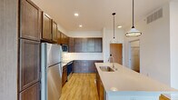 This thoughtfully designed single-floor residence combines modern finishes with practical living spaces throughout. The kitchen serves as a central highlight, showcasing rich dark wood cabinetry, light quartz countertops, a classic white subway tile backsplash, and a full suite of stainless steel appliances including a refrigerator, microwave, and range. A generous island with an undermount sink and stylish pendant lighting anchors the space, offering both prep space and a natural gathering point. Ample upper and lower cabinetry extends along one wall, providing generous storage. The living area opens to a private balcony through a sliding glass door, with transom windows above allowing additional natural light and scenic outdoor views of greenery and water. The bedrooms feature plush carpeting and large black-framed windows that frame picturesque natural views, filling each room with abundant daylight. Additional rooms with light wood-look flooring offer flexibility for various uses. The bathroom is equipped with accessibility grab bars, a white vanity with quartz countertop, and a framed mirror. A dedicated laundry area includes a stacked Whirlpool washer and dryer alongside built-in cabinetry with a folding surface, adding everyday convenience to this well-considered layout.