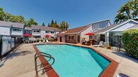 Located at 395 Ano Nuevo Ave in Sunnyvale, California, this amenity space serves as the indoor spa and hot tub facility for a residential complex. The compact, single-floor interior is defined by clean white walls, tile flooring, and a standout vaulted ceiling equipped with dual skylights that flood the room with natural light. At the heart of the space sits a brick-trimmed spa and hot tub, finished with blue and red accent tiles and accessible via stainless steel handrails. A large open doorway connects the indoor spa directly to the outdoor pool area, creating a seamless indoor-outdoor experience. Outside, the community showcases a generously sized swimming pool with a distinctive angular shape, surrounded by a spacious concrete sundeck furnished with lounge chairs and red patio umbrellas. Mature trees provide natural shade around the perimeter, while a secured wrought iron fence encloses the entire pool area for privacy and safety. Adjacent to the pool deck, a fitness center and additional seating areas round out the amenities. The surrounding residential buildings feature classic gabled rooflines and multi-level layouts, creating a welcoming neighborhood atmosphere under the sunny Sunnyvale skies.