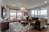Located in the heart of Kansas City's vibrant Westport neighborhood, this single-floor rooftop lounge offers a sophisticated and welcoming communal space. The interior design blends mid-century modern aesthetics with contemporary finishes, centered around warm hardwood flooring that runs throughout the open layout. A large, tufted olive-green sectional sofa anchors the main seating area, complemented by cognac leather accent chairs arranged around a round walnut coffee table atop a geometric patterned area rug. A striking capiz shell globe pendant light hangs overhead, adding an elegant focal point to the room. Textured vertical-stripe wallcovering lines the walls, lending depth and warmth to the space. Oversized black-framed floor-to-ceiling windows flood the interior with natural light and frame sweeping panoramic views of the Kansas City skyline. A wall-mounted flat-screen television is positioned opposite the main seating arrangement for comfortable viewing. A built-in wet bar features navy cabinetry, dark countertops, and a dramatic dark tile backsplash, providing a functional service area. Floor-to-ceiling sliding glass doors open directly onto a furnished outdoor terrace, complete with seating and a grill, seamlessly extending the lounge experience to the outdoors.