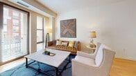 Located at 1200 N Garfield St in Arlington, Virginia, this single-floor residence offers a thoughtfully designed layout with clean lines and contemporary finishes throughout. The open-concept kitchen is a standout feature, equipped with stainless steel appliances including a gas range, built-in microwave, and refrigerator, complemented by warm wood cabinetry, dark granite countertops, and a tile mosaic backsplash. A central island with bar seating and pendant lighting above serves as both a functional workspace and a casual dining area, with light hardwood flooring tying the space together seamlessly. The living room is bright and welcoming, with large windows that invite natural light and provide outdoor views, creating an airy ambiance ideal for relaxing or entertaining. The bedrooms offer a serene retreat, each featuring neutral tones, soft carpeting, and windows that allow ample natural light to filter in. The bathroom is finished with large-format tile flooring, a full tub-shower combination with clean white tile surround, and a wood-accented vanity with an integrated sink. Throughout the residence, the consistent use of warm wood tones, neutral palettes, and modern fixtures creates a cohesive and inviting atmosphere well-suited for comfortable urban living in the heart of Arlington.