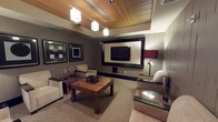 This elegantly appointed single-floor space exudes a refined hospitality ambiance, thoughtfully designed for comfort and style. The ceiling is a standout architectural feature, showcasing a warm slatted wood design accented by recessed lighting panels and a row of modern pendant lights that cast a soft, inviting glow throughout the room. The walls are finished in a muted taupe tone, providing a neutral backdrop that complements the rich wood and upholstered furnishings. A large wall-mounted flat-screen television is set within a bold, oversized dark frame, serving as the focal point of the main seating area. Flanking the TV are framed black-and-white wall art pieces that add a touch of contemporary sophistication. The seating arrangement features plush, neutral-toned lounge chairs positioned around a substantial wood-grain coffee table, creating an intimate and functional gathering space. A secondary accent table with a dark glass top is positioned nearby, accompanied by a stylish red-shaded lamp that introduces a subtle pop of color. The carpeted floor adds warmth underfoot, while the overall layout maximizes the sense of space. The cohesive design blends modern and transitional elements, making this a polished and welcoming environment suited for relaxation or private gatherings.