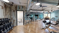 This two-floor fitness center at 1180 Cushing Circle in Saint Paul, Minnesota, offers a well-appointed exercise facility designed to support a comprehensive workout experience. The upper level serves as the primary cardio floor, featuring a striking curved ceiling with recessed lighting and ceiling fans. Large floor-to-ceiling windows flood the space with natural light, complementing the signature teal-painted walls and warm hardwood-style flooring. Rows of treadmills, ellipticals, and stationary bikes are thoughtfully arranged throughout. The lower level transitions into a strength and free-weights area, distinguished by its industrial-chic aesthetic — exposed ductwork, pendant lighting, and ceiling fans pair with a distinctive reclaimed wood accent wall in muted blues, grays, and browns. This floor houses a robust selection of strength machines, free weights, a cable system, and dedicated lifting equipment. Two staircases connect the floors for easy access. The facility also includes two accessible bathrooms — one with a roll-in shower — as well as a sauna, providing a full recovery amenity. Full-length mirrors throughout the strength area enhance the sense of space. The overall design balances modern industrial character with a welcoming, energetic atmosphere.
