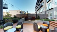 This outdoor garden space offers a well-designed terrace that blends modern aesthetics with natural elements to create a comfortable and inviting open-air environment. The warm-toned wood deck serves as the foundation of the space, providing a clean and contemporary surface that anchors the overall layout.

A standout feature of the garden is a rectangular water fountain set into a raised concrete surround, creating a gentle cascading water effect that adds both visual interest and a calming ambient sound to the space. Surrounding plantings of shrubs, ornamental grasses, and perennials line the perimeter, softening the hardscape and adding natural texture and seasonal color.

Seating areas are arranged throughout the deck, featuring dark wicker lounge chairs and round stools accented with bright yellow cushions, complemented by low glass-top tables. The arrangement encourages social gathering and relaxed outdoor use. String lights with globe-style pendants are strung overhead, providing soft evening illumination and adding a warm, inviting atmosphere after dark.

A horizontal-slatted privacy fence defines the garden boundary, while lush plantings along the building's base further integrate the architecture into the landscape. The open sky above ensures abundant natural light throughout the day, making this a functional and attractive outdoor retreat.