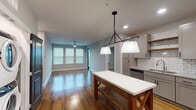 This single-floor residence in Rogers, Arkansas offers a thoughtfully designed layout with contemporary finishes throughout. The open-concept living and kitchen area is anchored by warm hardwood flooring that reflects natural light streaming in through multiple windows fitted with blinds. A ceiling fan overhead and recessed lighting ensure the space remains well-lit at all hours.

The kitchen is a standout feature, outfitted with gray shaker-style cabinetry, a classic white subway tile backsplash, granite-style countertops, and a full suite of stainless steel appliances including a refrigerator, range, microwave, and dishwasher. Open floating shelves add a modern touch, while a freestanding island with a white top and rustic wood base serves as a functional centerpiece beneath a striking black pendant light fixture.

The bedrooms feature plush carpet flooring, ceiling fans, and natural light from windows. Dark-panel interior doors add a bold contrast against the light gray walls throughout. A generous walk-in closet equipped with multi-tier wire shelving provides ample storage.

The two full bathrooms include gray vanities with stone-look countertops, large mirrors, and tub-shower combinations—one featuring large-format marble-style tile surround. In-unit laundry is also present, adding everyday convenience to this well-appointed home.
