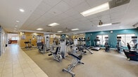 Located at 6503 Marsol Rd in Mayfield Heights, Ohio, this well-appointed fitness center offers a generously sized, single-floor layout designed to support a full range of exercise needs. The main exercise room is expansive and thoughtfully organized, with distinct zones for both strength training and cardiovascular activity. The strength training area is equipped with multiple selectorized weight machines, a cable cross system, free-weight dumbbells on a tiered rack, and adjustable benches — providing options for a comprehensive full-body workout. The cardio zone features rows of treadmills, elliptical trainers, and stationary bikes, each equipped with individual media screens for an engaging workout experience. Large wall mirrors along one side of the room amplify the sense of space and allow users to monitor their form. The flooring transitions from durable carpet tiles in the exercise areas to clean, large-format ceramic tile in the entryway and hallway, creating a practical and polished aesthetic. Warm tan and teal accent walls add visual interest, while recessed lighting and ceiling fans ensure a bright, comfortable environment. Natural light filters in through multiple windows fitted with blinds. A clean, functional bathroom completes the facility, rounding out this well-equipped fitness space.