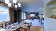 This single-floor space combines classic design elements with a warm, inviting atmosphere throughout its open-concept layout. The living room serves as a central gathering area, featuring plush wall-to-wall carpeting and a generously sized L-shaped sectional sofa in a rich dusty rose hue, accented with gold and earth-toned throw pillows. A plaid upholstered armchair and an oval glass-top coffee table complete the seating arrangement. The room benefits from abundant natural light streaming through multiple windows dressed with elegant layered drapery featuring dark valances and neutral sheers. French doors open directly to an outdoor deck or patio, offering views of surrounding greenery. Crown molding adds a refined architectural detail along the ceiling line. The adjacent dining area is defined by warm hardwood flooring and anchored by a dark wood dining table set with formal place settings. An ornate wrought-iron chandelier overhead adds character to the space. A dark wood china cabinet provides both display and storage. The seamless flow between the dining and living areas creates a cohesive, functional space ideal for both everyday living and entertaining.