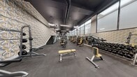 Located at 18 Lane Ave in Caldwell, NJ, this professionally designed fitness facility spans two floors, offering a comprehensive and well-appointed workout environment. The upper level — the larger of the two floors — serves as the primary strength training floor, featuring a broad, open layout lined with commercial-grade machines accented in yellow and grey, adjustable benches, a full dumbbell rack, and a dedicated functional training zone complete with an artificial turf area, kettlebells, stability balls, and battle ropes. Floor-to-ceiling windows along one wall flood the space with natural light, while a dark ceiling with exposed ductwork and stylish pendant lighting lend an industrial-modern aesthetic. A bold geometric-patterned accent wall adds a distinctive visual element near the interconnecting staircase, which features glass railings and clean metallic finishes. The lower level houses dedicated cardio and group cycling studios, each enclosed with glass partitions for an open yet defined feel. A row of treadmills and ellipticals lines one studio, while the cycling room is outfitted with a fleet of stationary bikes. Mirrored walls throughout both levels amplify the sense of space and support training visibility. The dark tile flooring, black-finished ceilings, and consistent modern design language create a cohesive, high-energy atmosphere throughout the entire facility.