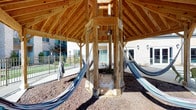 The Hammock Pavilion is a thoughtfully designed outdoor amenity space that combines rustic charm with comfortable leisure. At its centerpiece stands a robust timber-framed pavilion with an impressive pitched roof constructed from warm natural wood beams, creating an open-air retreat with ample shade and ventilation. Multiple fabric hammocks are suspended throughout the structure, offering inviting spots to relax, while a swing adds an additional element of casual recreation. A decorative stone base anchors the central support column, adding a refined architectural touch to the natural aesthetic.

Surrounding the pavilion, a gravel ground cover complements the organic feel of the structure, and the space opens to views of adjacent buildings, a pool area, and landscaped grounds. Nearby, an interior gathering space extends the amenities, featuring a spacious lounge with a dark tile fireplace, track lighting, ceiling fans, and comfortable seating arrangements. A dining area with high-top seating, a kitchenette with warm cabinetry, and large windows and glass doors overlooking the pool and courtyard complete the indoor component. Together, the pavilion and interior spaces create a well-rounded, community-focused environment designed for both active enjoyment and relaxed socializing.