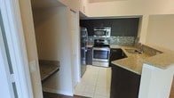 This single-floor space showcases a thoughtfully designed kitchen as its focal point. The kitchen is finished with rich, dark espresso-toned cabinetry that extends to the upper and lower levels, providing ample storage. Stainless steel appliances, including a French door refrigerator, over-the-range microwave, and electric range, lend a sleek, contemporary feel. The countertops are finished in granite with warm gold and brown tones, complementing the cabinetry beautifully. A mosaic tile backsplash in neutral tones of grey and brown adds texture and visual interest to the cooking area. The kitchen floor is laid with light-toned ceramic tile, creating a bright contrast against the darker cabinetry. A peninsula with matching granite extends into the adjacent area, offering additional counter space and a natural transition to the living area. Adjacent to the kitchen, a change in flooring to warm hardwood tones suggests a distinct living or dining zone. A closeted storage area is conveniently located near the kitchen, adding to the unit's practical layout. Overall, the space presents a modern, functional design with quality finishes throughout.