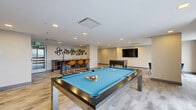 Located at 880 Pacific St in Stamford, Connecticut, this generously sized game and entertainment lounge offers a well-appointed, multi-zone layout designed for recreation and socializing. The space features light wood-tone flooring throughout, recessed lighting, and a clean, contemporary aesthetic with neutral walls accented by a bold deep-blue feature wall in the lounge area.

At the heart of the game room sits a sleek billiards table with a blue felt surface, complemented by a foosball table nearby. A wet bar with dark countertops and hexagonal wall décor serves as a social hub, flanked by modern orange bar stools. A wall-mounted flat-screen television provides an additional entertainment focal point, set against a gray accent wall with built-in display niches.

The lounge zone offers comfortable seating, including tan leather armchairs and neutral sofas arranged around wooden side tables, ideal for relaxed gatherings. A separate area with a long conference-style table and office chairs adds a functional, collaborative dimension to the space.

Large floor-to-ceiling windows and glass sliding partitions flood the interior with natural light while framing views of the surrounding Stamford cityscape. The open, flowing layout connects each zone seamlessly, creating a versatile environment suited for both active recreation and casual lounging.