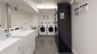 This shared laundry facility is designed for practicality and ease of use, offering a clean and well-organized environment for residents. The room features a combination of front-loading and top-loading washing machines alongside matching dryers, providing ample capacity for multiple users. A dedicated payment kiosk is conveniently located near the entrance, streamlining the laundry process. A utility sink and a white folding counter offer additional functionality for hand-washing and sorting garments. The flooring consists of durable grey tiles, complementing the bright white walls and ceiling that keep the space feeling fresh and hygienic. Fluorescent strip lighting ensures the room is well-lit throughout. Ventilation ductwork is visible along the upper walls, supporting efficient appliance operation. Posted instructional signage assists users with proper machine operation. The room is accessed through a half-glazed door fitted with a ventilation grille, allowing for airflow while maintaining security. A tall dark storage unit near the entrance provides additional organizational space. Overall, this laundry room is a functional, well-maintained shared amenity designed to accommodate the everyday needs of building residents with efficiency and convenience.