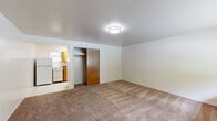 This single-floor unit offers a straightforward, functional layout designed to make efficient use of its available space. The main living area is finished with wall-to-wall neutral carpeting underfoot and white walls throughout, creating a clean, understated backdrop. A flush-mount ceiling light provides overhead illumination, while a window along the front wall admits natural light and offers a view of surrounding greenery. A front entry door opens directly into this open living space, which also features a floor vent for climate control.

Adjacent to the living area, a carpeted room serves as the sleeping space, equipped with a sliding wood-panel closet that provides convenient built-in storage. The room mirrors the neutral palette of the rest of the unit, maintaining a simple and cohesive aesthetic.

The galley-style kitchen runs along one wall and is finished with light tile flooring in a classic diamond pattern. It includes warm-toned wood cabinetry above and below the counter, a stainless steel sink, a granite-style laminate countertop, and white appliances including an electric range and refrigerator.

The bathroom continues the tile flooring found in the kitchen and features a wood-toned vanity with a granite-style countertop, a toilet, and a towel bar, all enclosed by a solid wood door.