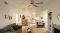 This single-floor space is defined by its notably high ceilings, which lend a sense of openness and airiness throughout. A large ceiling fan anchors the living area, enhancing both the visual height and comfort of the room. The living space features a plush sofa, a low-profile coffee table, and a decorative console table along one wall, creating a comfortable and functional arrangement. Neutral, light-toned carpet flows throughout, contributing to the warm and cohesive aesthetic.

The open layout connects the living area directly to a casual dining space, which is furnished with a wooden table and matching chairs alongside a hutch for additional storage. Natural light filters in through a doorway or window at the far end, brightening the dining nook. The kitchen sits adjacent to the dining area and features light wood cabinetry, countertop appliances including a microwave, and a compact, efficient layout. The cream and beige color palette runs consistently throughout all areas, creating a unified, inviting ambiance suited to everyday living.