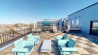 This expansive rooftop terrace is a thoughtfully designed outdoor amenity space that combines style, comfort, and breathtaking surroundings. The deck is finished with a combination of wood-tile flooring in both warm and gray tones, creating distinct zones across the generous open-air layout. The exterior walls of the rooftop structure are clad in blue horizontal lap siding, lending the space a coastal, resort-inspired aesthetic.

Seating throughout the terrace is eclectic and inviting, featuring mint-green perforated metal chairs and tables, vibrant coral and pink benches, woven rattan egg chairs suspended for a relaxed lounging experience, and wicker accent chairs with bold orange cushions. A wall-mounted outdoor flat-screen television is positioned along the blue-sided structure, ideal for outdoor entertainment. A full-length mirror and wall-mounted lighting fixtures further enhance the space's functionality.

The terrace commands sweeping panoramic views in multiple directions, including expansive suburban and city skylines backed by distant mountain ranges visible on clear days. The open railing design maximizes sightlines and ensures an unobstructed outdoor experience under open skies. Wall-mounted sconce lighting ensures the space remains welcoming into the evening hours, making this rooftop terrace a versatile destination for relaxation and social gatherings alike.