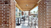 This covered outdoor patio offers a comfortable and functional space for socializing and relaxation. The structure is defined by exposed natural wood beam ceilings supported by substantial brick columns, creating a warm, semi-open-air environment that provides shade while maintaining an open connection to the outdoors. The concrete floor extends throughout the patio, providing a clean, durable surface underfoot.

The space is furnished with multiple high-top bistro-style tables paired with slatted patio chairs, arranged to accommodate both small and larger groups. The seating arrangement encourages casual gatherings while allowing for easy reconfiguration. Overhead lighting and cable infrastructure are integrated into the wooden ceiling, ensuring the space remains functional into the evening hours.

Brick walls frame the perimeter, giving the patio a grounded, architectural character that complements the surrounding buildings visible in the background. Lush landscaping, blooming shrubs, and mature palm trees are visible beyond the patio's open edge, contributing to a pleasant outdoor ambiance. The overall setting is bright, airy, and well-suited for outdoor dining or leisure.