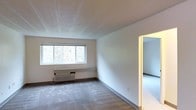 This single-floor space presents a clean, functional layout with a straightforward design. The main living area features neutral white walls and wall-to-wall carpeting in a soft gray tone, creating a quiet, understated atmosphere. A wide, multi-pane window dominates one wall, drawing in natural light and offering a view of surrounding greenery, contributing a sense of openness to the interior. Beneath the window sits a wall-mounted air conditioning unit, providing in-unit climate control. The flat, paneled ceiling maintains a modest height consistent with the room's practical character. A doorway leads directly into an adjacent room, also carpeted, suggesting a connected and accessible floor plan. The overall aesthetic is minimal and neutral, offering a blank canvas well-suited for personalization. White baseboards provide a clean border between the walls and flooring throughout. The space is designed with simplicity in mind, prioritizing functionality and ease of use. The natural light filtering through the main window enhances the brightness of the interior, making the space feel welcoming despite its understated design.