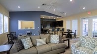 This single-story space showcases an open-concept layout that seamlessly connects the living, dining, and kitchen areas into one cohesive environment. The vaulted ceilings add a generous sense of vertical space, while recessed lighting and a ceiling fan ensure both bright illumination and comfortable airflow throughout.

The living area is anchored by a large, neutral-toned sofa accented with decorative pillows in blue and grey patterns, complemented by a dark wood coffee table and coordinating accent chairs. A bold slate-blue accent wall adds depth and visual interest, contrasting attractively with the warm beige walls throughout the rest of the space.

The kitchen features dark espresso cabinetry, black appliances, and a raised breakfast bar with blue upholstered bar stools. A pass-through window connects the kitchen to an adjacent area, enhancing the open feel. Multiple dining sets with dark wood chairs are positioned throughout the space, offering flexible seating arrangements. Natural light filters in through window treatments on one side, adding warmth to the overall atmosphere.