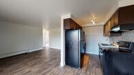 This single-floor residence offers a well-organized layout that balances functional living spaces with practical design. The open living area is a standout feature, showcasing durable wood-look flooring that extends throughout the main areas and a striking floor-to-ceiling stone fireplace that serves as a focal point on one wall. A sliding glass door adjacent to the fireplace allows natural light to filter in and provides access to an outdoor area. A wall-mounted air conditioning unit and baseboard heating ensure year-round comfort.

The galley-style kitchen is efficiently designed, featuring dark espresso cabinetry with sleek hardware, light countertops, a mosaic tile backsplash, a deep stainless steel sink, and black appliances including a full-size refrigerator and range. Track lighting overhead adds a modern touch to the space.

The bedrooms are carpeted for comfort and feature neutral tones, baseboard heating, and windows that bring in natural light. At least one bedroom includes a double closet with built-in hanging rods, offering generous storage. A hallway connects the living areas to the bedrooms and a full bathroom, which includes a shower-tub combination, toilet, vanity, and wood-look flooring with a small window for ventilation.