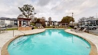 Located at 9200 N Normandale St in Fort Worth, Texas, this outdoor amenity area centers around a generously sized freeform swimming pool with a distinctive figure-eight silhouette. The pool's vivid turquoise water is complemented by a broad, stone-tiled deck that wraps around the entire perimeter, providing ample space for relaxation and recreation. Depth markers are clearly visible along the pool's edge, ensuring safety for all users.

The deck is appointed with multiple lounge chairs and seating arrangements, offering residents comfortable spots to unwind. A pergola structure with a stone base accent wall sits adjacent to the pool, providing a shaded retreat ideal for gathering. Additional pergola structures are positioned around the deck, further enhancing the outdoor living experience.

The area is enclosed by a black iron fence that defines the space while maintaining open sightlines to the surrounding mature trees and neighboring buildings. Exterior lighting poles are strategically placed throughout the area to ensure visibility after dark. Adjacent to the pool, a clubhouse or amenity building with large glass windows and a gabled roof serves as an architectural anchor for the space, offering indoor-outdoor connectivity. The overall design creates an inviting, functional outdoor environment suited for both leisure and community activity.
