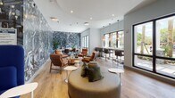 The AMLI Inspiration Room at 8021 Peters Rd in Plantation, Florida, is a thoughtfully designed communal amenity space that seamlessly blends creative aesthetics with functional versatility. The single-floor layout features light wood flooring throughout, contributing to a warm and cohesive atmosphere across all zones.

A striking full-wall mural dominates a large portion of the space, featuring an intricate, abstract composition in moody blues, grays, and whites that serves as both a bold artistic statement and a dynamic backdrop for the various seating areas. This mural wraps into the dedicated co-working zone, where a long wooden conference table is surrounded by ergonomic rolling chairs and complemented by cognac leather lounge chairs — ideal for both focused work sessions and informal meetings.

The lounge area is anchored by a large, rounded sectional sofa in soft gray, paired with cognac leather chairs and minimalist white accent tables, creating an inviting setting for casual gathering. High-back, cobalt blue privacy chairs offer quieter nooks for individual focus or one-on-one conversations.

A built-in bar counter runs along a wall of large, black-framed windows, which flood the space with natural light and provide views of the surrounding greenery. Copper pendant lights hang above the counter, adding warmth and a refined industrial touch. The overall ambiance is modern, energetic, and professionally curated.