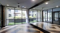 This single-floor studio space is thoughtfully designed to create an open, energizing environment ideal for fitness, yoga, or movement-based activities. The most striking architectural feature is the expansive floor-to-ceiling glass wall system framed in sleek black metal, which floods the interior with abundant natural light and offers unobstructed views of a well-maintained outdoor courtyard complete with green lawn, manicured hedges, and a pergola structure beyond. A full-length mirrored wall runs along one side of the room, amplifying the sense of openness and allowing for form monitoring during exercise. The flooring is a standout design element, featuring a bold herringbone-patterned wood floor that adds warmth and visual interest to the otherwise clean, contemporary aesthetic. Recessed ceiling lighting complements the natural light, while a ceiling fan ensures comfortable airflow throughout the space. The dark-framed architectural accents — from the window mullions to the ceiling beams — create a modern industrial contrast against the bright, airy interior. Overall, the space strikes a balance between functionality and refined design, offering a bright, welcoming environment suitable for a variety of active uses.