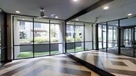 This single-floor studio space is thoughtfully designed to create an open, energizing environment ideal for fitness, yoga, or movement-based activities. The most striking architectural feature is the expansive floor-to-ceiling glass wall system framed in sleek black metal, which floods the interior with abundant natural light and offers unobstructed views of a well-maintained outdoor courtyard complete with green lawn, manicured hedges, and a pergola structure beyond. A full-length mirrored wall runs along one side of the room, amplifying the sense of openness and allowing for form monitoring during exercise. The flooring is a standout design element, featuring a bold herringbone-patterned wood floor that adds warmth and visual interest to the otherwise clean, contemporary aesthetic. Recessed ceiling lighting complements the natural light, while a ceiling fan ensures comfortable airflow throughout the space. The dark-framed architectural accents — from the window mullions to the ceiling beams — create a modern industrial contrast against the bright, airy interior. Overall, the space strikes a balance between functionality and refined design, offering a bright, welcoming environment suitable for a variety of active uses.