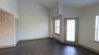 This two-story space is defined by its striking vaulted ceilings that rise dramatically overhead, creating an open and airy atmosphere. The main living area showcases rich, dark hardwood-style flooring that provides a bold contrast against the neutral, light-toned walls throughout. Large windows fitted with horizontal blinds allow ample natural light to fill the space, enhancing its bright and welcoming feel. A glass-panel exterior door leads to what appears to be a balcony or outdoor area, further extending the sense of openness and connectivity with the outdoors. The clean, minimalist architectural design is complemented by simple baseboards and subtle wall detailing, offering a versatile canvas for a variety of interior styles. The elevated ceiling height in the main area adds a sense of volume and grandeur rarely found in standard multi-floor layouts. The upper level is suggested by the architectural lines of the vaulted structure, contributing to the multi-dimensional character of the space. Overall, the combination of soaring ceilings, natural light, and quality flooring gives this space a modern, polished ambiance that is both functional and visually impressive.