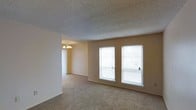 This single-floor residential space offers a clean, functional layout designed for comfortable everyday living. The main living area is a standout feature, showcasing two large windows fitted with horizontal blinds that allow generous natural light to fill the room, creating a bright and welcoming atmosphere. The walls are finished in a warm neutral tone, complementing the soft, wall-to-wall carpeting that extends throughout the space and adds a cozy underfoot feel. The textured popcorn ceiling adds a classic touch consistent with traditional residential construction. Adjacent to the living area, a dining space is visible, distinguished by an elegant chandelier with multiple bulbs that provides warm, ambient lighting. A set of vertical blinds nearby hints at additional access to the outdoors or further natural light. The open flow between the living and dining areas enhances the sense of space and makes the layout feel connected and versatile. Overall, the space presents a straightforward and practical residential environment with neutral finishes, ample natural light, and a seamless transition between functional zones.