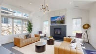This contemporary community clubhouse offers a thoughtfully designed, open-plan gathering space spread across a generous single-level footprint. The main lounge is anchored by a sleek, dark-surround fireplace with a mounted display above, flanked by large windows that fill the room with natural light and frame views of the surrounding neighborhood. Soaring vaulted ceilings accented by a modern chandelier lend an airy, expansive feel to the seating area, which features comfortable leather sofas arranged around a round coffee table on a light herringbone area rug. A built-in bar counter with gold-toned stools lines one wall, offering a casual spot for socializing. The entryway is bright and welcoming, with floor-to-ceiling glass doors that open directly to the outdoor amenity area, where a sparkling pool, a fire pit lounge with navy blue seating, and a spacious concrete patio create an ideal setting for outdoor gatherings. Back inside, a well-appointed kitchenette is outfitted with white shaker cabinetry, frosted glass upper cabinets, a penny-tile backsplash, gray quartz countertops, hexagonal tile flooring, a stainless steel double sink, a built-in microwave, and a dishwasher. Throughout, crisp white walls and abundant natural light create a clean, modern ambiance.