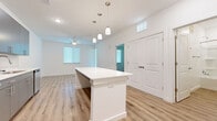 This thoughtfully designed one-bedroom, one-bathroom residence offers a clean, modern aesthetic across a single floor. The open-concept living and kitchen area serves as the heart of the home, featuring warm luxury vinyl plank flooring, crisp white walls, and recessed lighting that creates a bright, welcoming atmosphere. A substantial white kitchen island anchors the space, illuminated by a trio of pendant lights hanging overhead. The kitchen is well-appointed with sleek gray cabinetry, a white countertop, subway tile backsplash, and a full suite of stainless steel appliances, including a refrigerator, range, microwave, and dishwasher. The living area extends naturally from the kitchen, enhanced by a ceiling fan and multiple windows fitted with blinds that allow for natural light control. The bedroom offers a comfortable retreat, finished with plush carpeting, a ceiling fan, and a window with a view of greenery outside. A dedicated closet provides convenient storage. The bathroom maintains the home's clean design language with light wood-look flooring, a full soaking tub with shower, a dark-toned vanity, and bright white walls and fixtures throughout.