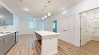 This thoughtfully designed one-bedroom, one-bathroom residence offers a clean, modern aesthetic across a single floor. The open-concept living and kitchen area serves as the heart of the home, featuring warm luxury vinyl plank flooring, crisp white walls, and recessed lighting that creates a bright, welcoming atmosphere. A substantial white kitchen island anchors the space, illuminated by a trio of pendant lights hanging overhead. The kitchen is well-appointed with sleek gray cabinetry, a white countertop, subway tile backsplash, and a full suite of stainless steel appliances, including a refrigerator, range, microwave, and dishwasher. The living area extends naturally from the kitchen, enhanced by a ceiling fan and multiple windows fitted with blinds that allow for natural light control. The bedroom offers a comfortable retreat, finished with plush carpeting, a ceiling fan, and a window with a view of greenery outside. A dedicated closet provides convenient storage. The bathroom maintains the home's clean design language with light wood-look flooring, a full soaking tub with shower, a dark-toned vanity, and bright white walls and fixtures throughout.