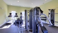 This fitness center offers a thorough and well-organized workout environment designed to accommodate a wide range of exercise needs. The space is outfitted with a robust selection of cardio machines, including treadmills and ellipticals, positioned near large windows that allow natural light to fill the room and create an energizing atmosphere. At the center of the facility, a multi-station cable and strength training machine serves as a focal point, providing users with versatile options for resistance and muscle-building workouts. Along the perimeter, a fully stocked dumbbell rack offers an extensive range of free weights, catering to both beginner and advanced fitness levels. The flooring features durable, rubber-textured material suited for high-traffic athletic use, while recessed lighting in the ceiling supplements the natural light for consistent visibility throughout the space. The walls are painted in a soft, neutral green tone, contributing to a calm and focused workout environment. The overall layout is thoughtfully arranged to maximize usable floor space and allow for comfortable movement between equipment stations.