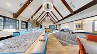 Located in Colorado Springs, CO, this Bear Run Point clubhouse is a thoughtfully designed community gathering space that combines mountain-inspired architecture with modern interior finishes. The single-floor layout offers generous square footage organized around a dramatic central great room anchored by soaring vaulted ceilings with exposed dark wood beam trusses and an array of statement pendant and ring chandelier fixtures that provide both ambient and accent lighting.

A floor-to-ceiling stacked stone fireplace with a mounted flat-screen television serves as the room's focal point, flanked by a comfortable sectional seating area atop herringbone-patterned hardwood flooring. A long granite-topped bar counter with cushioned high-back stools defines a casual lounge zone, while a separate dining area features bold, colorful geometric wall art paired with terracotta-upholstered chairs for a vibrant contrast.

Throughout the space, expansive black-framed glass doors and windows flood the interior with natural light and offer views to the outdoor surroundings. A quieter lounge area with tufted blue sectional sofas provides a relaxed retreat, separated by a central column. Patterned carpet tiles in muted blue-gray tones unify the various zones. The entry vestibule, with its large glass sliding doors, creates a bright and welcoming arrival experience.