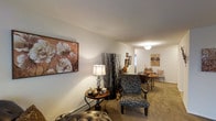This single-floor space presents a comfortable and well-appointed interior with a seamlessly connected living and dining area. The living area features wall-to-wall carpeting in warm neutral tones, providing a soft and inviting foundation. Smooth, light-colored walls create a clean backdrop that enhances the overall brightness of the space. A large floral canvas print adds a decorative focal point to one wall, complementing the warm, earthy color palette present throughout.

The living area is thoughtfully arranged with a patterned armchair and matching ottoman, alongside a classic round side table topped with a traditional table lamp, adding ambient warmth to the space. Decorative tall branch arrangements contribute an organic, stylized element to the room's aesthetic.

Transitioning toward the rear of the space, a dining area is visible, furnished with a dark metal dining set. A ceiling-mounted light fixture illuminates this area, providing functional overhead lighting. Additional framed artwork adorns the walls, maintaining a consistent decorative theme throughout. The open layout between the living and dining areas encourages an easy flow, making the space feel connected and functional.