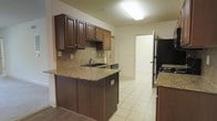This single-story space showcases a well-appointed kitchen as its focal point. The kitchen features rich, dark wood cabinetry with brushed nickel hardware, providing ample upper and lower storage. Granite countertops in warm earth tones offer generous prep space and extend across a peninsula that doubles as a breakfast bar or workspace. A tile backsplash in a neutral diagonal pattern complements the countertops and adds visual texture to the cooking area. Black appliances, including a refrigerator and dishwasher, provide a cohesive, modern contrast against the cabinetry. The flooring transitions between light-toned ceramic tile in the kitchen area and hardwood-style flooring nearby, clearly delineating functional zones. Overhead fluorescent lighting provides bright, even illumination throughout the kitchen. The open layout allows for easy movement between the kitchen and adjoining areas, with a doorway visible in the background offering seamless flow to other parts of the space. Neutral beige walls throughout contribute to a clean, understated aesthetic that allows the darker wood finishes to stand out as the primary design element.