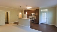 This single-floor space showcases a thoughtfully designed open-concept layout that seamlessly connects the kitchen and living areas. The kitchen is a standout feature, equipped with rich, dark wood cabinetry that extends to the upper walls, providing generous storage throughout. Stainless steel appliances, including a gas range and microwave, complement the sleek granite countertops that top both the cabinetry and the central peninsula. The peninsula serves as a natural divider between the kitchen and the adjoining living space, offering a functional surface for casual dining or additional prep space. Two pendant lights hang above the peninsula, adding focused task lighting and an elegant design accent, while a flush-mounted ceiling fixture provides broader illumination. Crown molding runs along the ceiling, lending a refined architectural detail to the space. The flooring transitions from warm, dark hardwood in the kitchen area to a lighter surface in the living space, creating a subtle visual distinction between zones. A door at the far end of the space allows for natural light to filter in. The overall ambiance is warm, modern, and functional, with a clean, well-appointed aesthetic throughout.