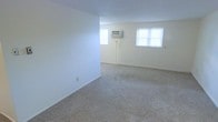 This single-floor space features an open main room finished with wall-to-wall beige carpeting and crisp white walls, creating a clean, neutral backdrop. Two windows allow natural light to filter in, brightening the interior and contributing to an airy atmosphere. A wall-mounted air conditioning unit is installed between the windows, providing climate control without occupying floor space. The room offers a generous, open layout with no interior partitions, making it highly adaptable for various furniture arrangements and uses. Baseboards run along the perimeter of the room, adding a simple, finished detail to the otherwise minimalist design. The overall ambiance is understated and functional, with a blank-canvas quality that allows for flexible customization of the living area.