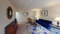 Located at 120 Patrick St SE in Vienna, Virginia, this single-floor residence offers a practical and comfortable layout designed for everyday living. The living room serves as the central gathering space, featuring plush carpeted flooring, neutral walls, and room for a full seating arrangement. A patterned area rug anchors the space, while wall-mounted artwork adds a decorative touch and ambient lighting creates a welcoming atmosphere. The galley-style kitchen is efficiently designed, offering warm wood cabinetry, stone-patterned countertops, a stainless steel sink, and black appliances including a gas range and refrigerator. A pass-through counter connects the kitchen to the adjacent living area, enhancing the open feel. The bedroom is a calm retreat with soft carpeting, a window with vertical blinds that allows natural light and views of the surrounding greenery, and ample space for furnishings. The full bathroom includes a pedestal sink, toilet, and a tub-shower combination with a curtain enclosure, finished in neutral tones for a clean, functional aesthetic. The overall floor plan, as seen in the dollhouse view, reflects a cohesive and well-proportioned single-level design suited for comfortable residential living in the heart of Vienna, VA.