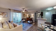 This single-floor residence offers a cohesive and thoughtfully designed layout that balances comfort with functionality. The living room serves as the heart of the home, featuring wood-style flooring, a ceiling fan with light fixture, and a large sliding glass door flanked by vertical blinds and drape panels that open to an outdoor balcony, filling the space with natural light and providing pleasant outdoor views. A peninsula counter connects the living area to the galley-style kitchen, which is outfitted with dark espresso cabinetry, light stone-look countertops, a mosaic tile backsplash, and black appliances including a refrigerator, electric range, and built-in microwave. The dining area accommodates a full-sized glass dining table beneath a multi-bulb chandelier, with a wall-mounted flat-screen frame adding a media element. Both bedrooms are carpeted and feature natural light from windows with blinds and decorative curtain panels. Two full bathrooms include dark wood vanity cabinetry, light countertops, framed mirrors with vanity lighting, and shower/tub combinations. Wood-style flooring carries through the bathrooms and main living areas, creating a consistent, modern aesthetic throughout the home.