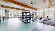 This expansive fitness center is designed to accommodate a full range of workout needs within a bright, open, and architecturally refined environment. The space is defined by soaring ceilings adorned with prominent wood beam accents and fitted with large ceiling fans, contributing to both the visual appeal and air circulation throughout. Rows of oversized windows line the walls on multiple sides, flooding the interior with natural light and offering views of the surrounding outdoor landscape, including palm trees and open grounds.

The flooring features a distinctive teal-toned surface with darker accent zones, providing designated areas for various workout activities. A large mirror wall reflects the space and enhances the sense of openness. Wall-mounted flat-screen televisions are positioned throughout for entertainment during workouts.

The strength training area is comprehensively outfitted with a multi-functional power rack system that supports TRX suspension training and a punching bag, a full dumbbell rack, multiple weight benches, and a cable machine. The cardio section features treadmills, elliptical trainers, and stationary cycling bikes. A battle rope and rubber matted zones provide dedicated areas for functional and high-intensity training. The overall ambiance is modern, clean, and energizing, reflecting a professional-grade fitness facility.
