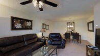 This single-floor residence offers a seamlessly connected living and dining area, creating an open and functional layout. The living space is anchored by rich, dark leather seating — a full sofa and matching armchair — arranged around a glass-top coffee table, lending a classic and polished aesthetic. Neutral-toned walls and light beige carpeting provide a clean, understated backdrop that complements the dark furnishings. A ceiling fan with an integrated light fixture overhead ensures both comfort and illumination in the living area. Framed wall art adds a decorative touch, contributing to the space's cohesive design. Transitioning naturally from the living area, the dining space is defined by a dark wood table and chairs, illuminated by an elegant pendant chandelier that adds a warm focal point. A decorative mirror on the dining wall enhances the sense of depth and brightness. The open flow between both areas makes the space feel inviting and well-proportioned, with consistent finishes and lighting choices tying the two zones together into a unified, comfortable living environment.