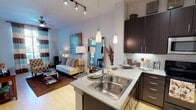 This single-story, one-bedroom, one-bathroom residence combines modern design with functional living. The open-concept kitchen is a standout feature, outfitted with dark espresso cabinetry extending to the ceiling, light granite countertops, a subway tile backsplash with under-cabinet lighting, stainless steel appliances, and a peninsula with a double stainless sink and bar seating illuminated by sleek pendant lights. Track lighting adds a contemporary touch overhead.

The living area flows naturally from the kitchen and is bathed in natural light from large windows dressed with striped curtains. A ceiling fan, light hardwood-style flooring, and a wall-mounted television contribute to the room's comfortable, modern ambiance. A sliding glass door leads to a private outdoor space.

The bedroom offers carpet flooring, a ceiling fan, natural light through a large window with outdoor views, and ample room for full-sized furniture. The bathroom features a full tub and shower combination with decorative tile accents and tile flooring.

A dedicated laundry area with a full-sized washer and dryer, plus built-in shelving, adds everyday convenience to this well-designed residence.